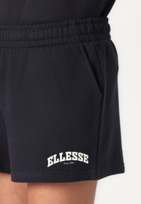 Tamno plave pamučne šortse s elastikom u struku, bočnim džepovima i bijelim logotipom "ELLESSE" blizu donjeg ruba. Glatka tekstura i opušteni kroj.
