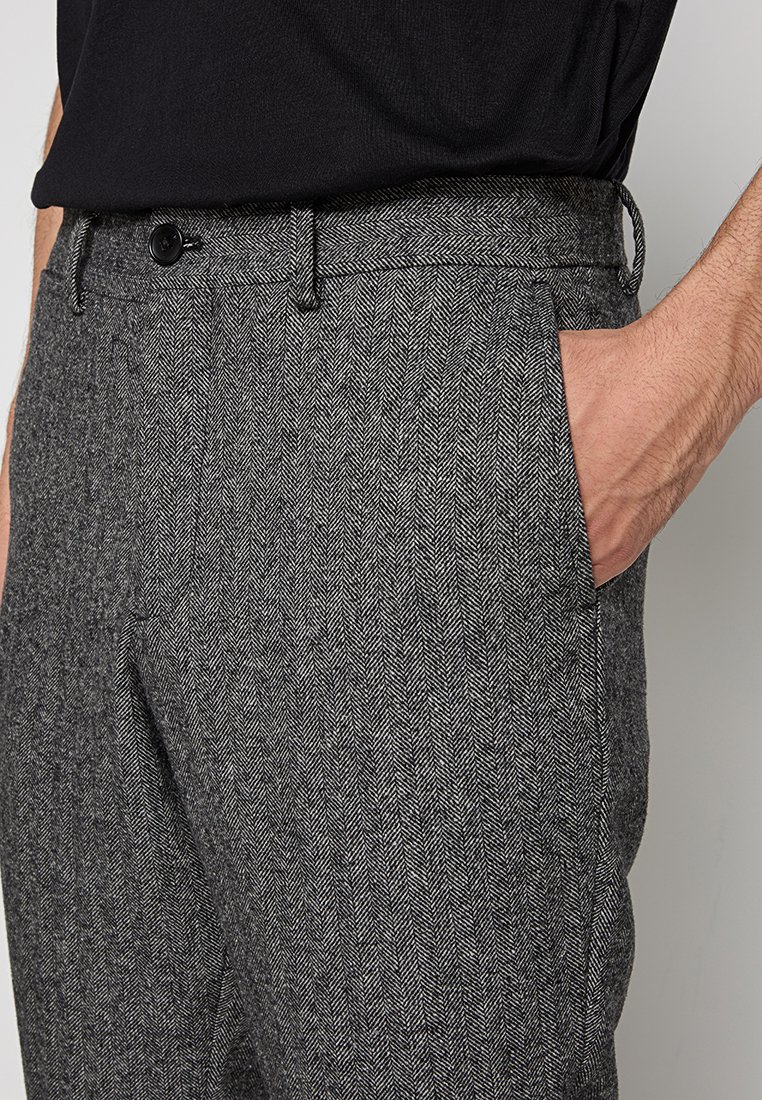 Pantaloni grigi a spina di pesce con una texture liscia, dotati di chiusura a bottone, passanti per cintura e tasche laterali. Vista ravvicinata della vita e della tasca.