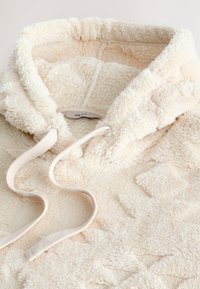 Hanorac crem din fleece sherpa, cu modele diamantate texturate și șnururi groase vizibile în zona gulerului.