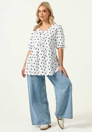 Blond kvinde iført en hvid skjorte med prikker og bindebånd samt lyseblå bukser med vide ben, poserer foran en ensfarvet baggrund.