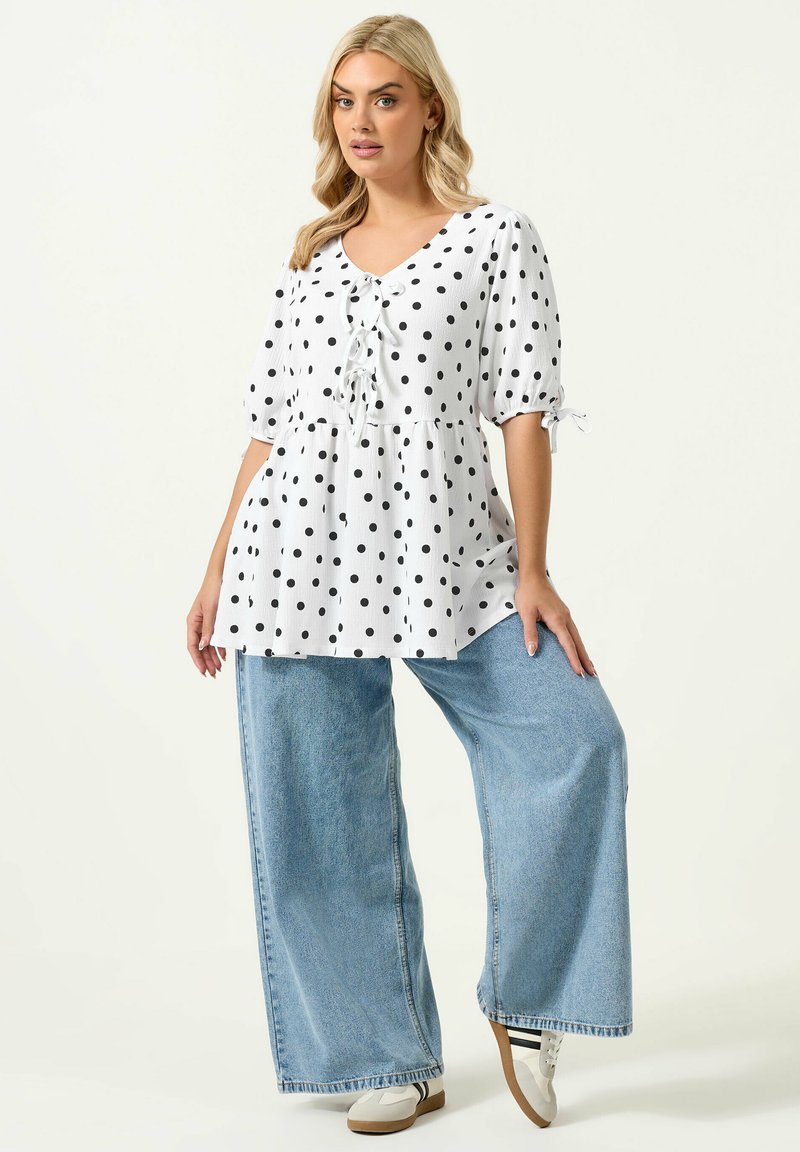 Blonde Frau, die eine weiße Bluse mit Punkten und Bindebändern sowie hellblaue Jeans mit weitem Bein trägt, posiert vor einem schlichten Hintergrund.