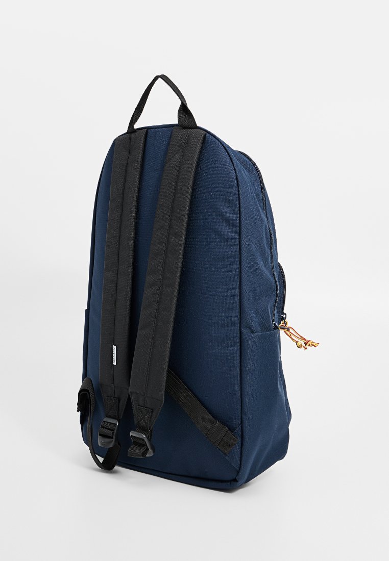 Sac à dos en tissu bleu marine avec des bretelles noires, forme arrondie, poche latérale et fermeture éclair avec un pompon décoratif. Surface texturée.