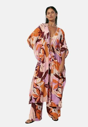 Kvinde iført løstsiddende, farverig kjole med blomsterprint og matchende bukser med vide ben og sandaler, poserende foran en ensfarvet hvid baggrund.