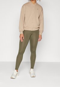 Felpa beige con bottoni e colletto rotondo, abbinata a leggings verde oliva e sneaker bianche. La felpa presenta un dettaglio con logo.