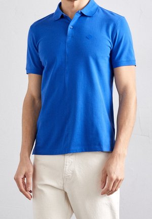 Blue polo shirt made of cotton, featuring a pointed collar, short sleeves, and a small logo accent on the chest. Paired with light-colored pants.