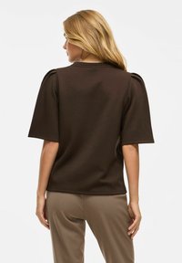 Brūns īsām piedurknēm tops ar puffed pleciem un apaļu kakla izgriezumu, izgatavots no mīksta auduma. Stilizēts ar beigu biksēm, rādot brīvu piegriezumu.