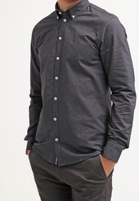 Chemise boutonnée gris foncé avec col, texture subtile et boutons blancs, associée à un pantalon foncé, mains dans les poches.