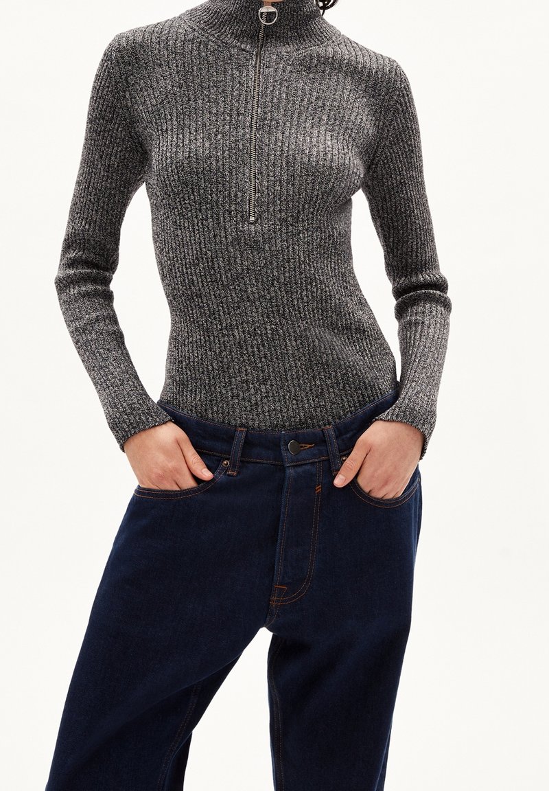Suéter de cuello alto acanalado en gris oscuro con un medio cierre en la parte delantera, combinado con jeans de mezclilla azul oscuro que tienen bolsillos y costuras de contraste.