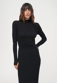Melnas garām piedurknēm tērptas turtlenecks, kas saskaņota ar ribotu melnu svārku. Audums izskatās gluds ar pieguļošu siluetu un šauriem piedurkņu galiem.
