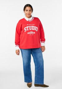 Červená mikina s bielym nápisom "STUDIO edition" v kombinácii s modrými džínsami. Biela golier s červenými pruhmi na výstrihu.