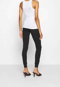 Bílý bezrukávový top a černé přiléhavé tepláky s žebrovanými cuffy a hladkým povrchem; model nosí černé lodičky na podpatku.