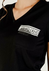 Czarny t-shirt z dekoltem w serek, z kieszonką na lewej piersi z białym i czarnym wzorem, ozdobioną ćwiekami i gładką teksturą.