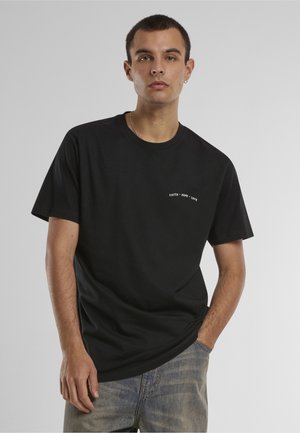 Jeune homme aux cheveux courts portant un T-shirt noir avec le texte « Foi • Espoir • Amour », posant avec une main dans la poche devant un fond gris.