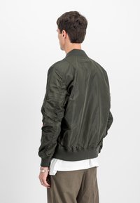Jachetă bomber verde cu guler și manșete ribbate. Prezintă o textură netedă și lucioasă, cu un design potrivit, evidențiind un tiv elastic.