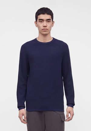Jeune homme aux cheveux courts noirs portant un pull texturé bleu foncé à manches longues et un pantalon gris avec une poche latérale, debout devant un fond uni.