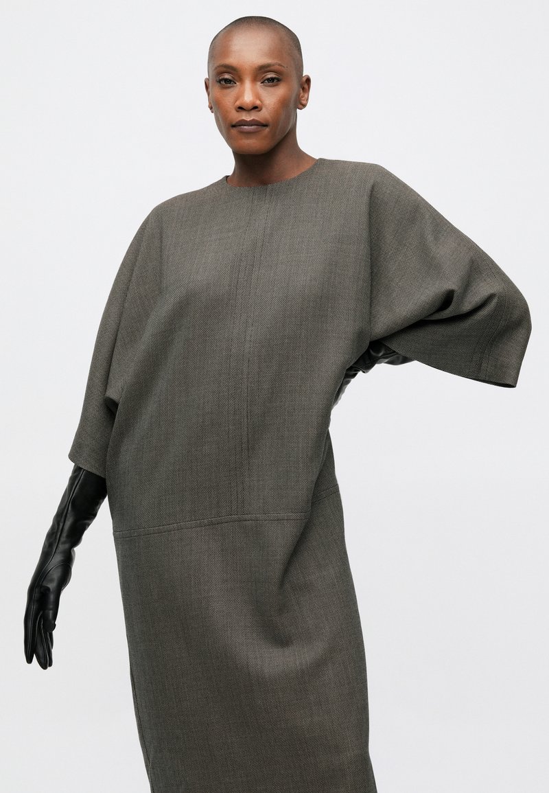 Femme avec la tête rasée portant une robe grise texturée ample et de longs gants noirs, posant contre un fond clair.