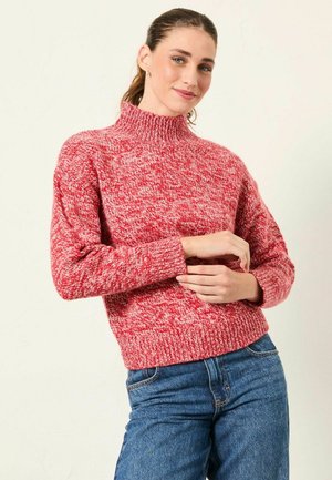 Rot-weißer, gesprenkelter Strickpullover mit hohem Rollkragen, langen Ärmeln und geripptem Saum, kombiniert mit blauen, hoch taillierten Jeans.