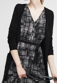 Cardigan in maglia nera sopra un vestito a V con motivo in bianco e nero. Il vestito presenta un design a griglia e una cintura in vita, con una texture liscia.