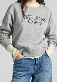 Šedá mikina s vyšívaným textem "PEPE JEANS LONDON" a květinovými akcenty. Žebrované manžety a lem. Pod ní je nošena krémová košile s dlouhým rukávem.