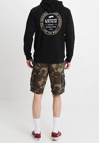 Felpa nera con cappuccio e logo Vans sulla schiena, abbinata a pantaloni cargo in camo. Calze bianche e sneakers rosse completano l'outfit.