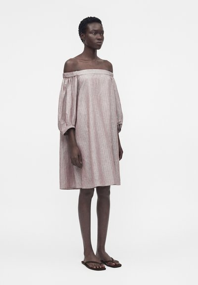 Γυναίκα που φοράει ένα φαρδύ, ροζ φόρεμα με ανοιχτούς ώμους και μανίκια τριών τετάρτων, καθώς και μαύρα σαγιονάρες, στέκεται μπροστά από ένα μονόχρωμο φόντο.