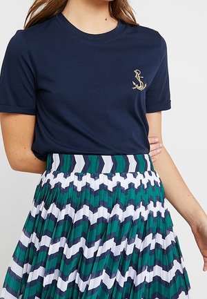 Camisa azul marino de manga corta con un pequeño bordado dorado de ancla combinada con una falda plisada con rayas en zigzag verde, blanca y azul marino.