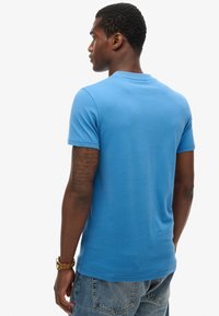 T-shirt in cotone blu con maniche corte, scollatura rotonda e una texture liscia; indossato con jeans in denim blu e un orologio d'oro.