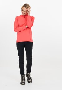 Korallenrotes Langarmshirt mit Reißverschlusskragen, kombiniert mit schwarzen, tapered Hosen und grauen Wanderschuhen. Das Shirt hat einen strukturierten Stoff und flache Nähte.