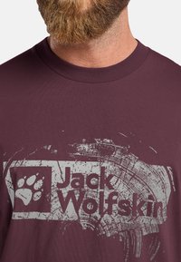 Bordeaux katoenen T-shirt met een versleten witte grafiek van een pootafdruk en de tekst "Jack Wolfskin" op de voorkant, ronde halslijn.
