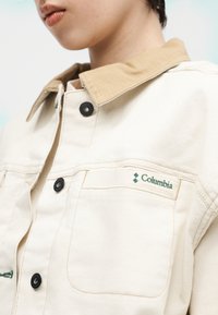 Μερική θέα ατόμου που φοράει μπεζ μπουφάν Columbia με γιακά σε αποχρώσεις καφέ και μαύρα κουμπιά, με το λογότυπο της μάρκας να εμφανίζεται στην τσέπη στο στήθος.