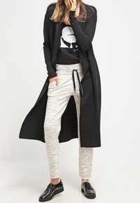 Persona che indossa un lungo cappotto nero, una t-shirt grafica nera, pantaloni grigi chiari con coulisse e scarpe lucide nere stringate.