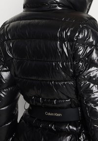 Chaqueta de plumas negra brillante con acolchado horizontal y un cinturón negro de Calvin Klein en la cintura, vista desde atrás.