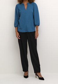 Blouse bleu sarcelle à manches bouffantes trois-quarts et petit col montant, associée à un pantalon noir  à jambes larges et à des talons noirs.