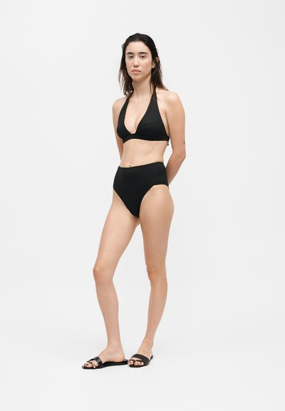 Γυναίκα που φοράει μαύρο μπικίνι με ψηλή μέση και μαύρα σανδάλια slide μπροστά σε λευκό φόντο.