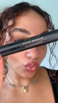 Personne aux cheveux bouclés tient un crayon pour yeux GLIDR noir étiqueté "Champagne Ice" près de son visage, les lèvres pincées, portant un collier cadenas doré.