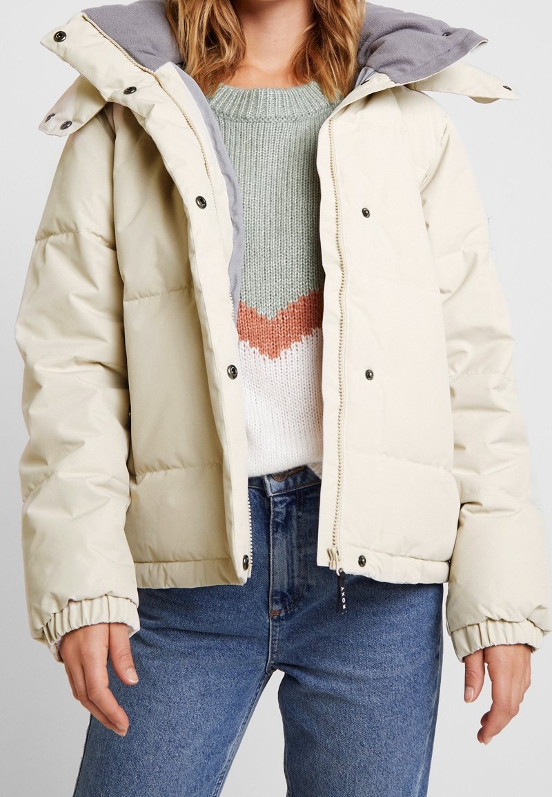 Femme portant une doudoune crème avec une capuche grise, un pull en tricot à motif chevron vert, blanc et orange, et un jean bleu.