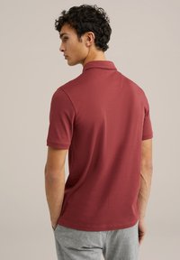 Camisa polo marrón con cuello y mangas cortas, que presenta una textura suave y detalles de costura sutiles. Combinada con pantalones gris claro.
