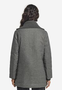 Gråullskappa med ribbad krage, med rak silhuett och långa ärmar. Texturerad tyg och minimal detaljer.