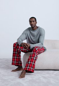 Pelēka, garām piedurknēm krekls, kas saskaņots ar sarkaniem un melniem pleda raksta biksēm. Audums izskatās mīksts. Models sēž uz gaiši krāsotas, teksturētas dīvāna.