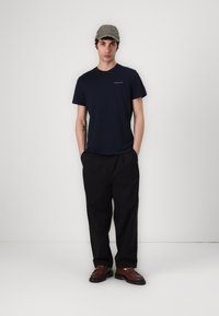 T-shirt navy à col rond avec un petit logo blanc, pantalon noir ample, et mocassins marron. Casquette vert olive avec détail brodé.