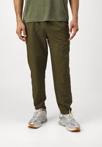 Olivenfarvede bukser med en tapered pasform, lavet af glat stof. Bæres med grå og sølv sneakers med en orange accent.