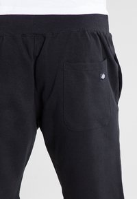 Gros plan sur un pantalon avec une ceinture élastique noire, une poche arrière rectangulaire et une petite étiquette de logo de marque sur la poche.