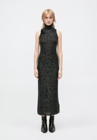 Black sleeveless turtleneck dress with a form-fitting silhouette, textured fabric featuring sparkling accents, paired with black ankle boots.