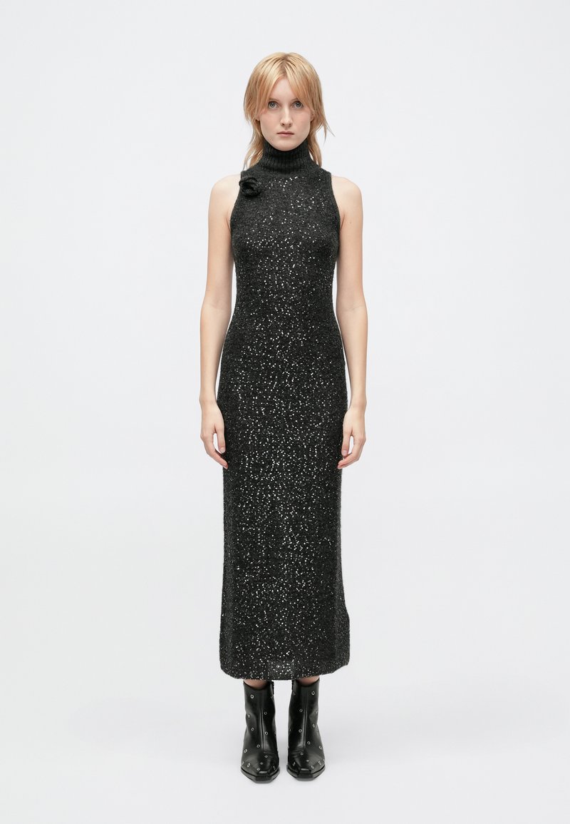 Black sleeveless turtleneck dress with a form-fitting silhouette, textured fabric featuring sparkling accents, paired with black ankle boots.