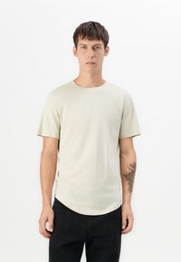 Camiseta beige clara hecha de una tela suave, cuello redondo, mangas cortas, dobladillo ligeramente curvado, usada con pantalones negros, mostrando un tatuaje visible en el brazo.