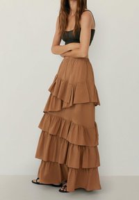 Jupe maxi marron à volants superposés, en tissu doux, taille élastique, longueur cheville, associée à un haut ajusté et des sandales plates.