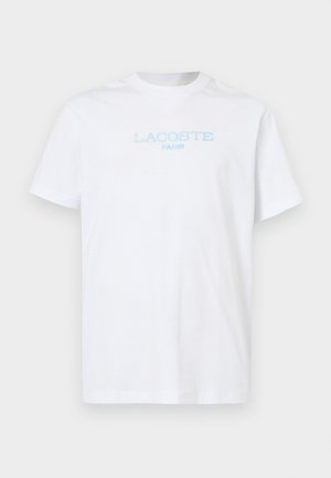 T-shirt en coton blanc à manches courtes et col rond. Présente un texte bleu clair "LACOSTE" et "PARIS" sur le devant.