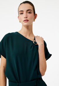 Teal bluse med draperet design, korte ærmer og en sølv cirkulær detalje på skulderen. Blødt stof.