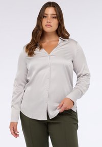 Blusa grigia chiara a maniche lunghe con scollatura a V, chiusura con bottoni e texture liscia. Indossata con pantaloni verde oliva.
