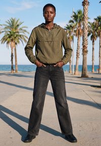 Mladić stoji sa rukama u džepovima na šetnici uz more obrubljenoj palmama pod vedrim nebom.